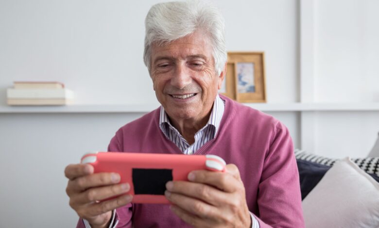 Elderly man smiling while playing on a handheld console indoors.