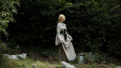 A cosplayer dressed as an elf in a fantasy costume stands in a lush, green forest.