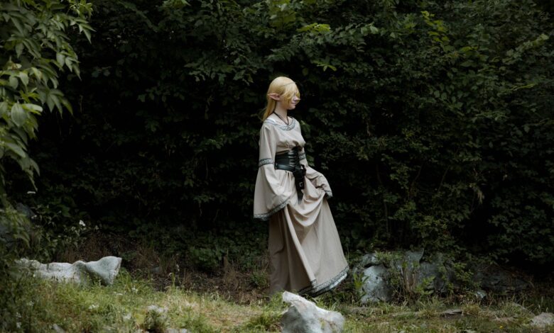 A cosplayer dressed as an elf in a fantasy costume stands in a lush, green forest.