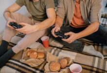 Two friends having fun playing video games with controllers and eating burgers indoors.
