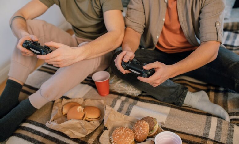 Two friends having fun playing video games with controllers and eating burgers indoors.
