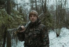 A man in camouflage holding a shotgun during winter hunting in the forest.