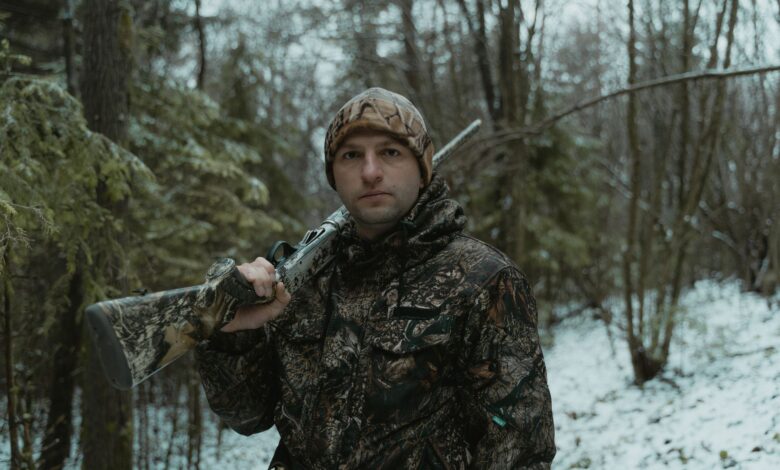 A man in camouflage holding a shotgun during winter hunting in the forest.