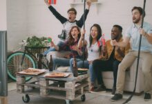 Excited friends cheering while watching a sports game on TV, holding flags and snacks.