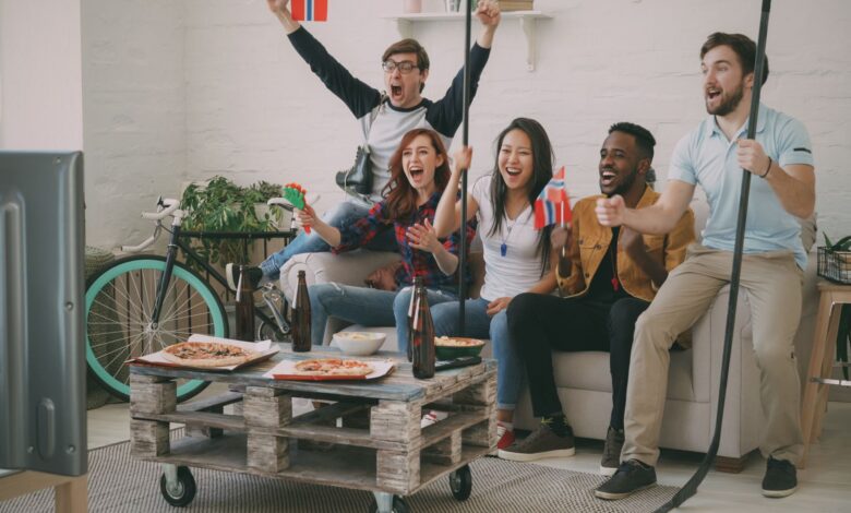 Excited friends cheering while watching a sports game on TV, holding flags and snacks.