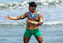 A tribal warrior cosplayer stands shirtless by the sea, wearing green shorts and golden armor parts.