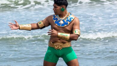 A tribal warrior cosplayer stands shirtless by the sea, wearing green shorts and golden armor parts.