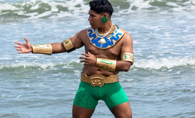 A tribal warrior cosplayer stands shirtless by the sea, wearing green shorts and golden armor parts.