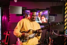 A man in a yellow hoodie plays a guitar simulation game indoors, immersed in gaming fun.