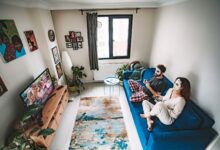 A couple playing video games on a couch with face masks, enjoying leisure time in a cozy living room.